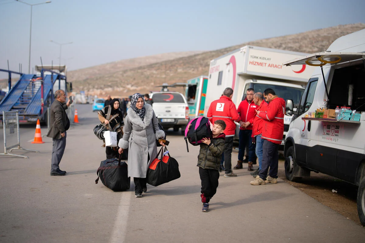 A family carries luggage along a road past vehicles and staff from the