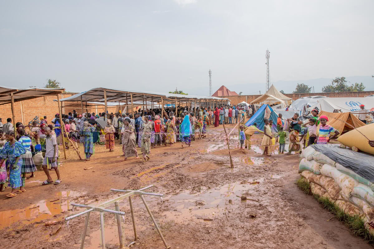 Large groups of people gather in the mud under makeshift shelters
