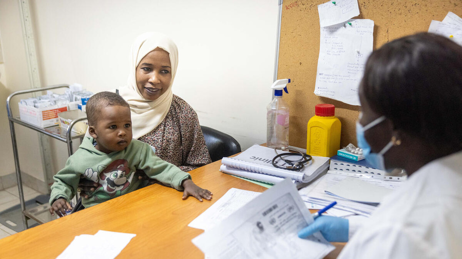 A woman is seated at a table with her young son, across from a medical staff member staff in a mask who is looking at paperwork